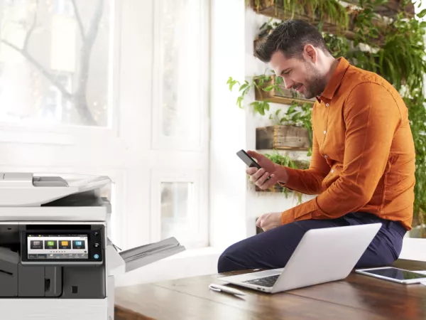 Man using laptop and mobile phone with a printer in foreground